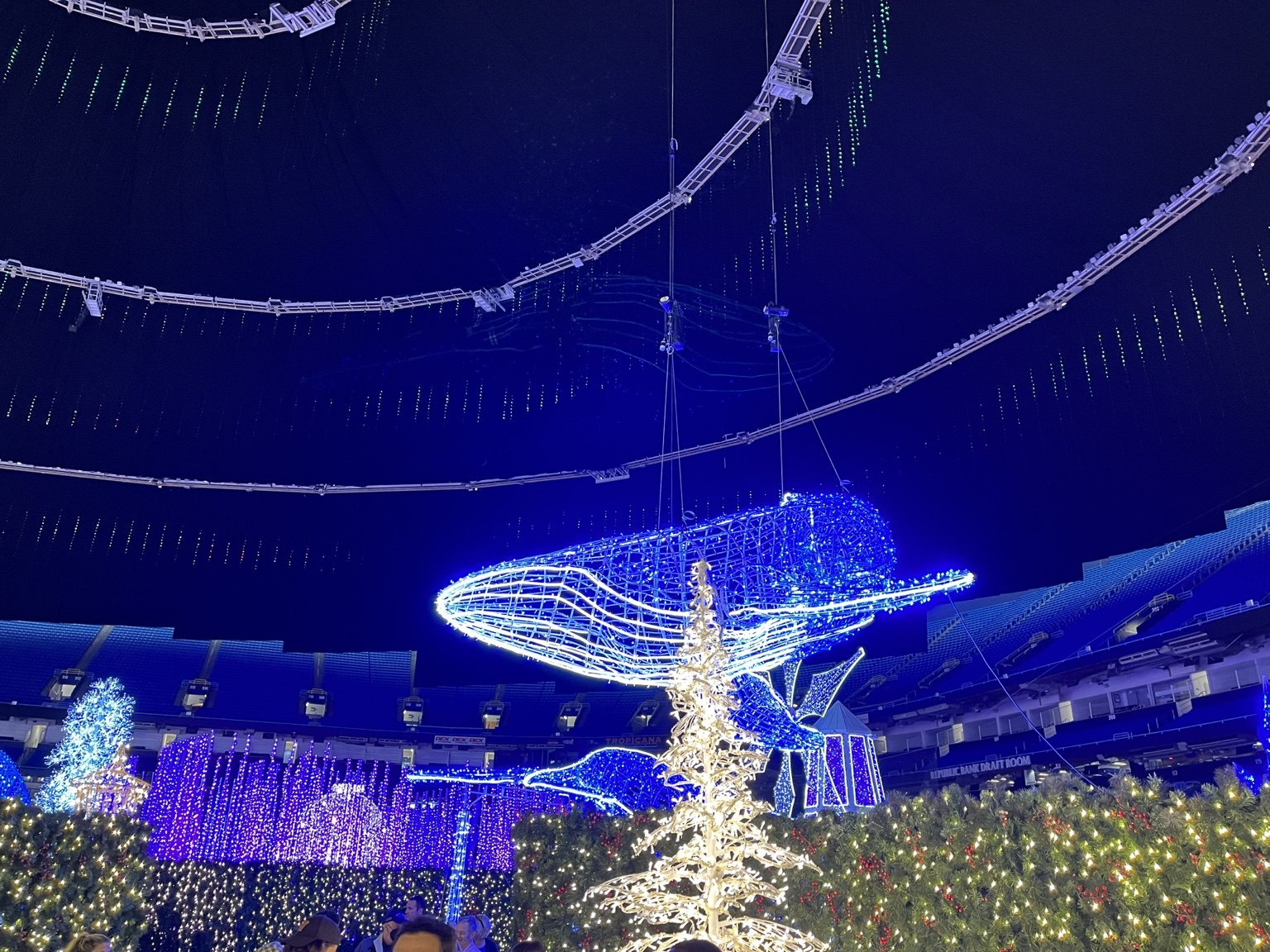 An illuminated blue whale flying above Christmas trees and light displays beneath the roof blackout